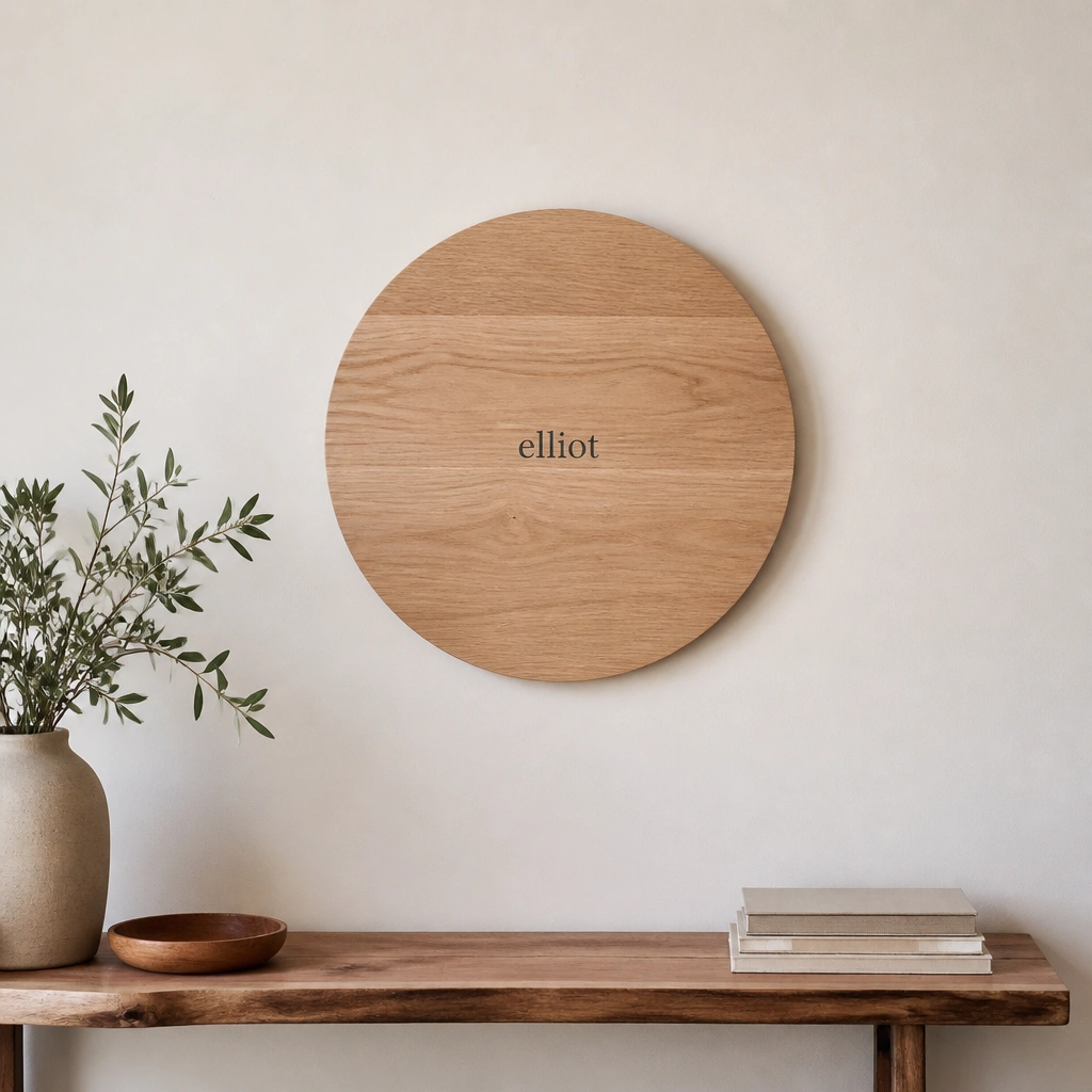 A round wooden disc mounted on a wall with a small centered name in dark gray vinyl. The wall is light and the disc is placed above a narrow console table.